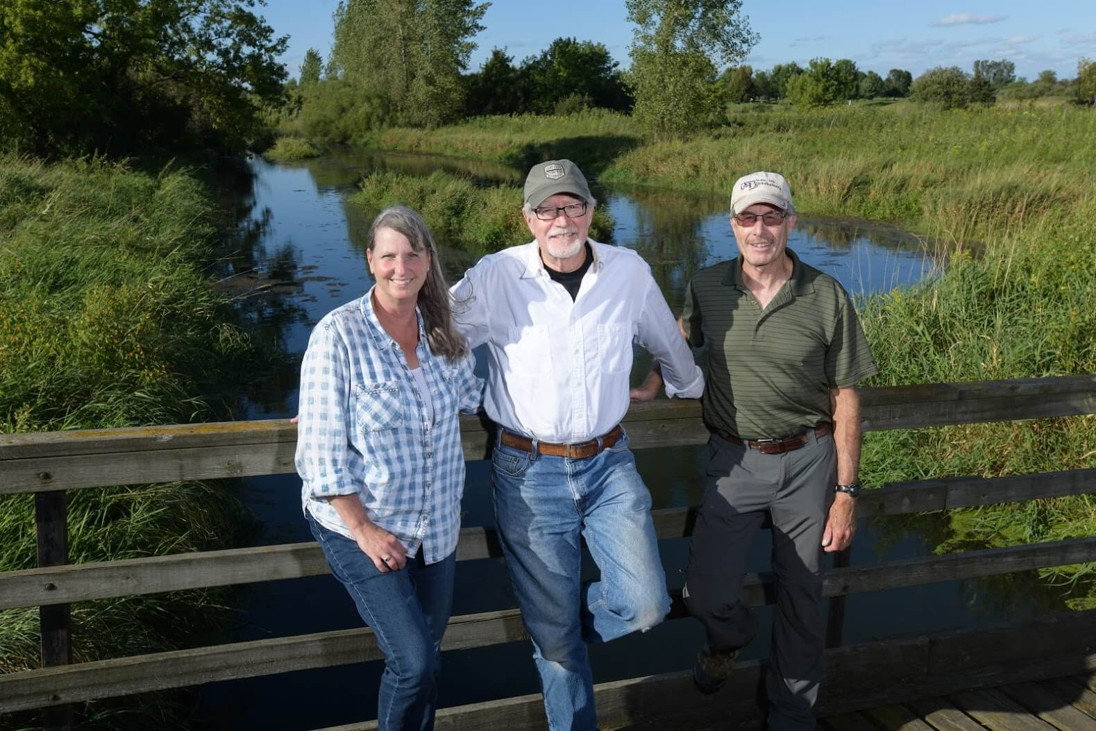 A woman and two men standing on a wooden bridge in front of wetlands.