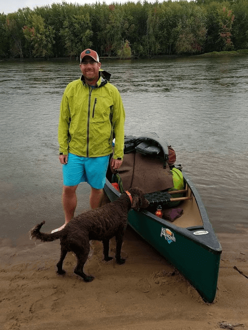 A middle aged man with a beard, yellow jacket, and blue shorts standing in shallow water next to a canoe and a brown dog.