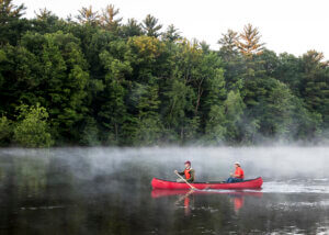 Two people paddling in a red canoe on the misty Wisconsin River, with a background of pine trees. The Wisconsin River has benefitted from the Knowles-Nelson Stewardship Program.