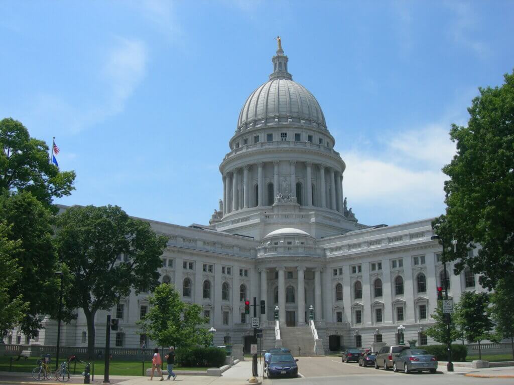 The Wisconsin State Capital building in summer with pedestrians and parked cars.