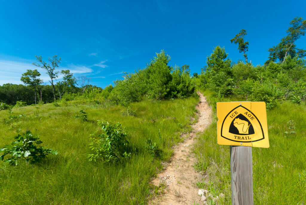 A small yellow sign along the Ice Age Trail with a narrow path leading into the woods.