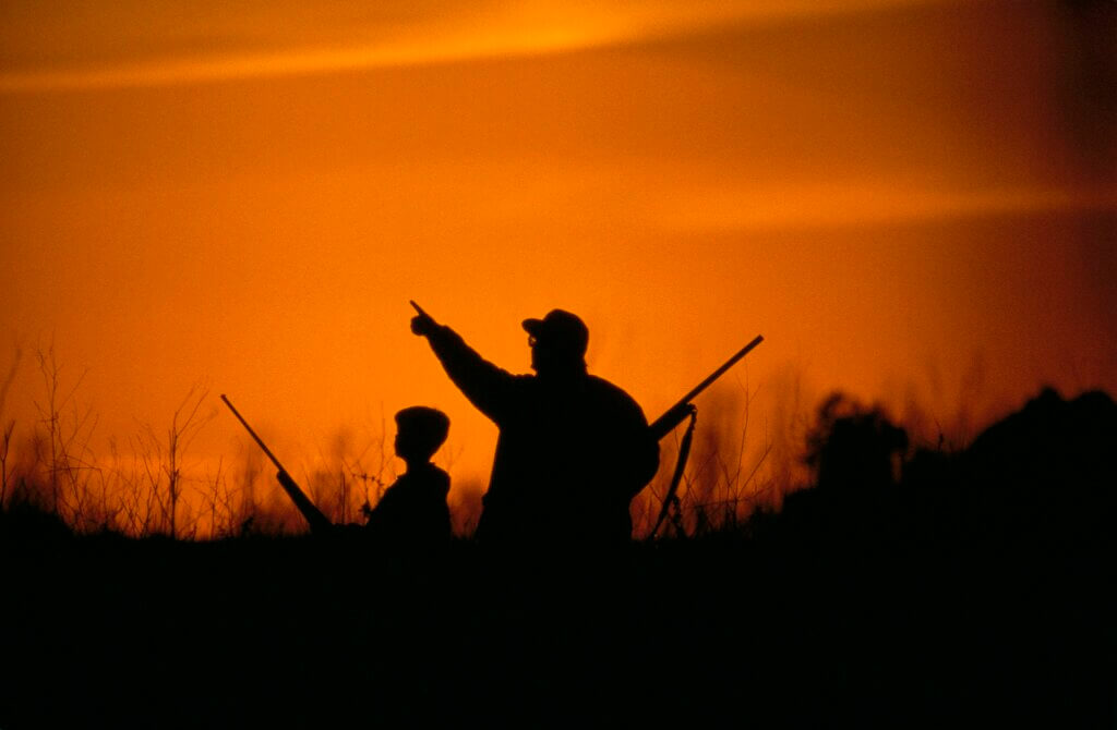Silhouettes of an adult and child duck hunting with a brilliant orange sky.
