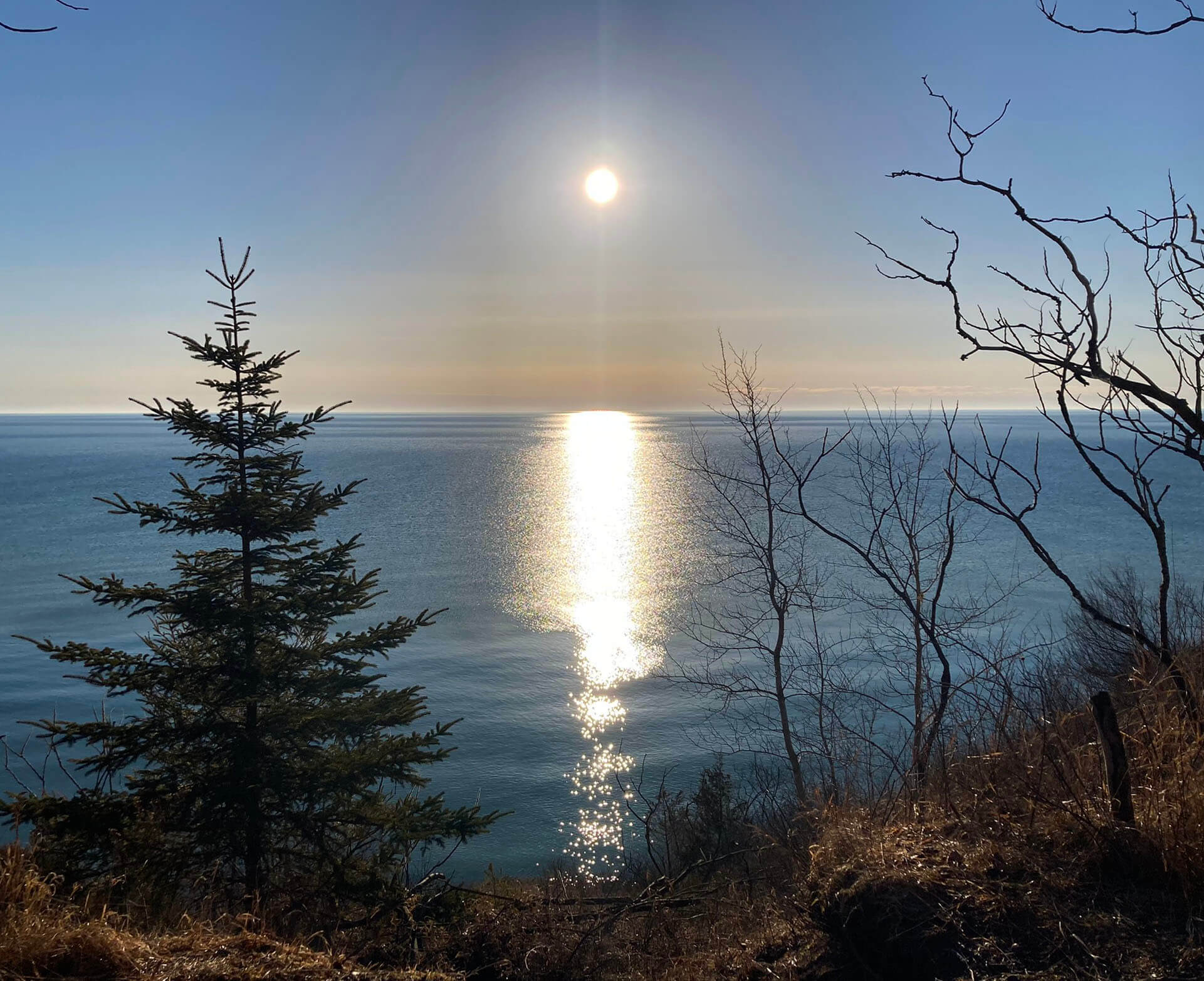 A late winter day at Lion's Den Gorge with the sun showing through a hazy sky over Lake Michigan and trees without any leaves along the shoreline.