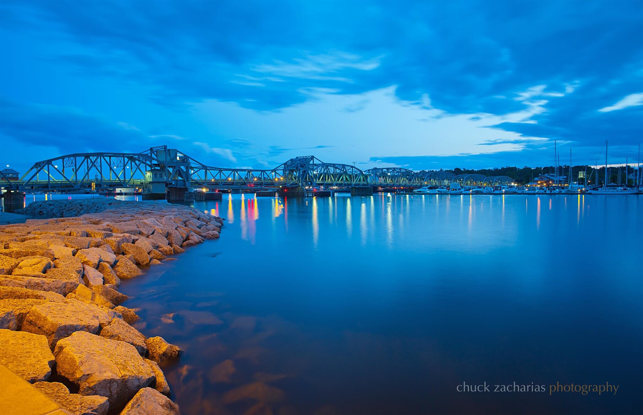 The Sturgeon Bay waterfront at twilight with the rocky shoreline on the left leading into a bridge over the water with anchored boats on the right.
