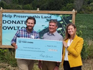 Knowles-Nelson Stewardship Program - Two men and a woman smiling and holding an oversized check for $2.3 million, standing in front of a sign about protecting the Cedar Gorge Clay Bluffs
