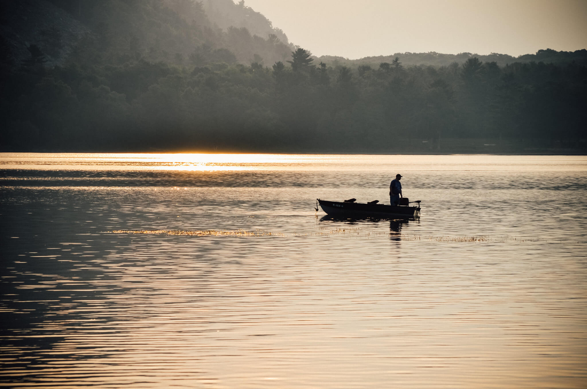 Knowles-Nelson Stewardship Program - a single fishing boat in the distance with a man standing and looking out at the water surrounded by dark gray trees and hills at dawn