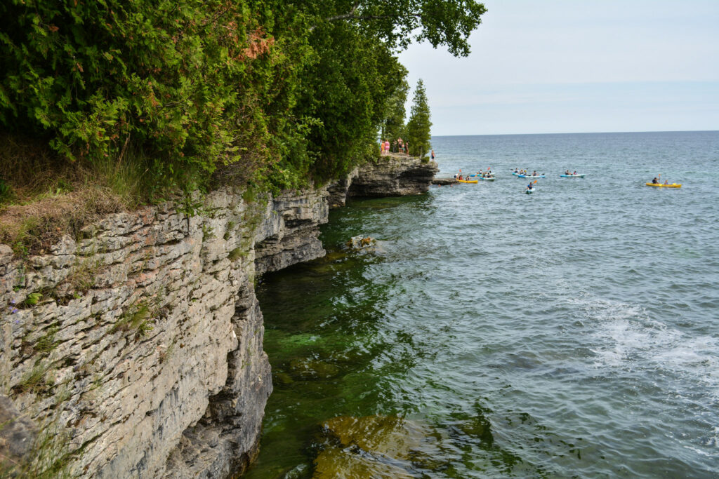 Water meets a steep outcropping with trees on top. There are several kayaks in the distance.