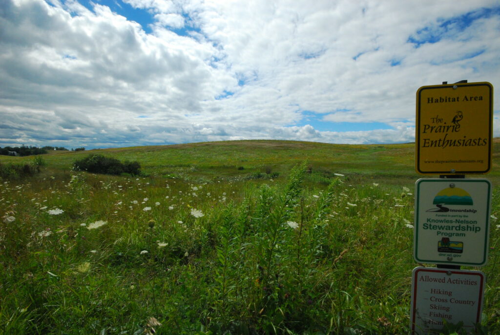 A rolling, green prairie with some white flowers. The sky is blue, with clouds. There is post with three information signs about the property.