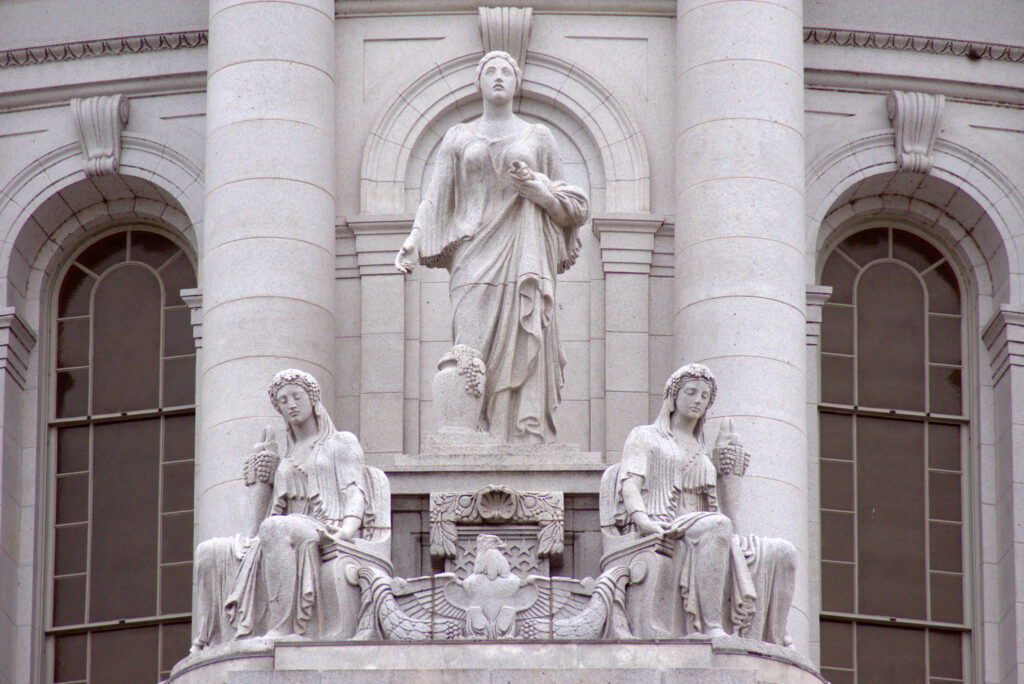 Close-up view of the exterior of the Wisconsin State Capitol building. Three stone statues of women are posed on a ledge in front of pillars.