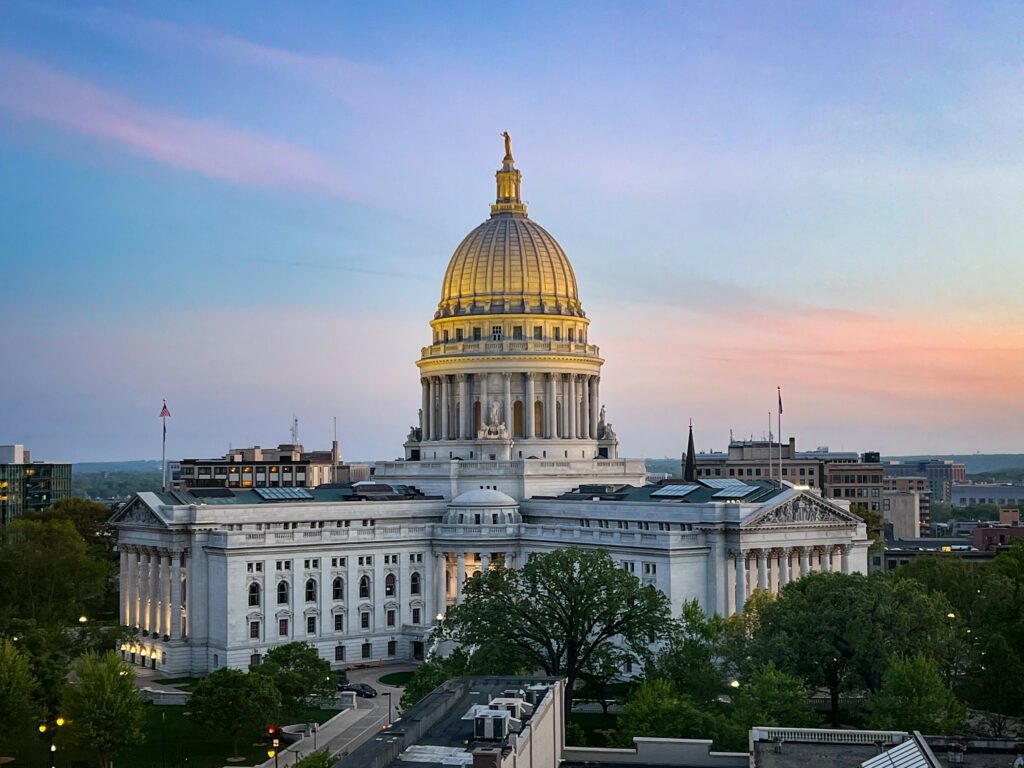 An aerial view of the Wisconsin Capitol building, illuminated by orange and pink light from a sunset.