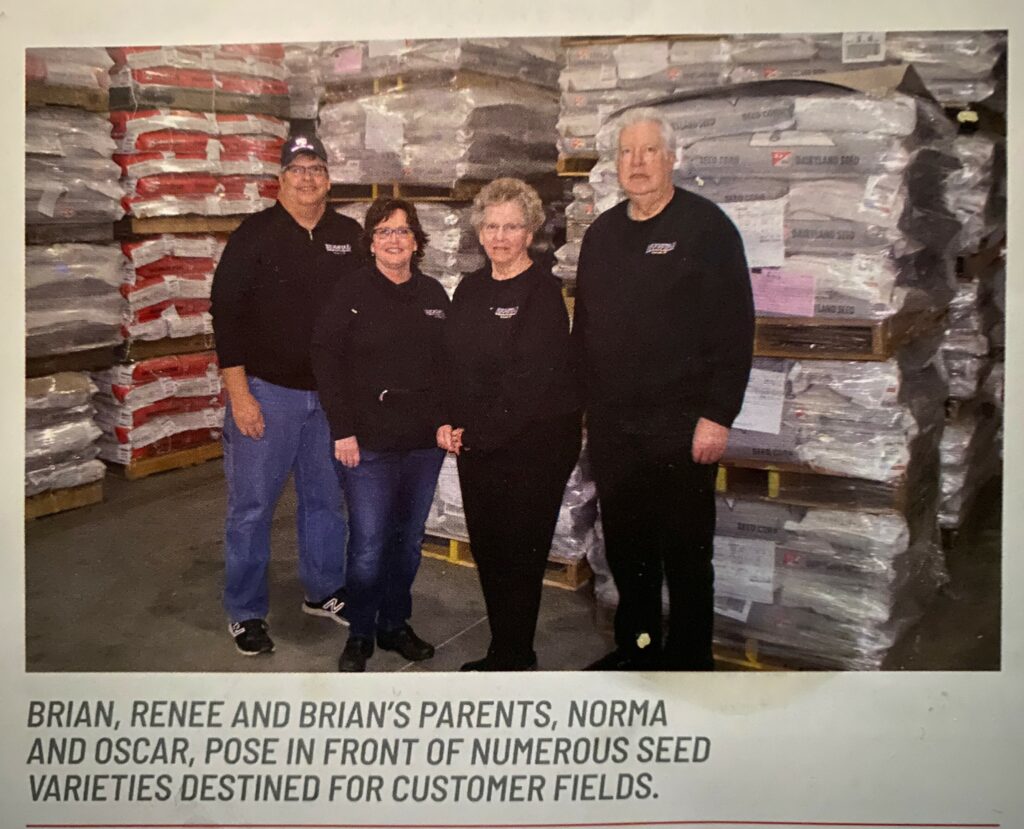 Four adults stand in front of pallets in a warehouse that are loaded with seed.