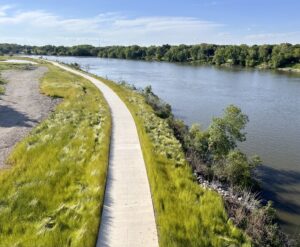 An aerial view of a paved path running along a river. There are trees on the opposite of the river, and the path is bordered with tall green grasses.