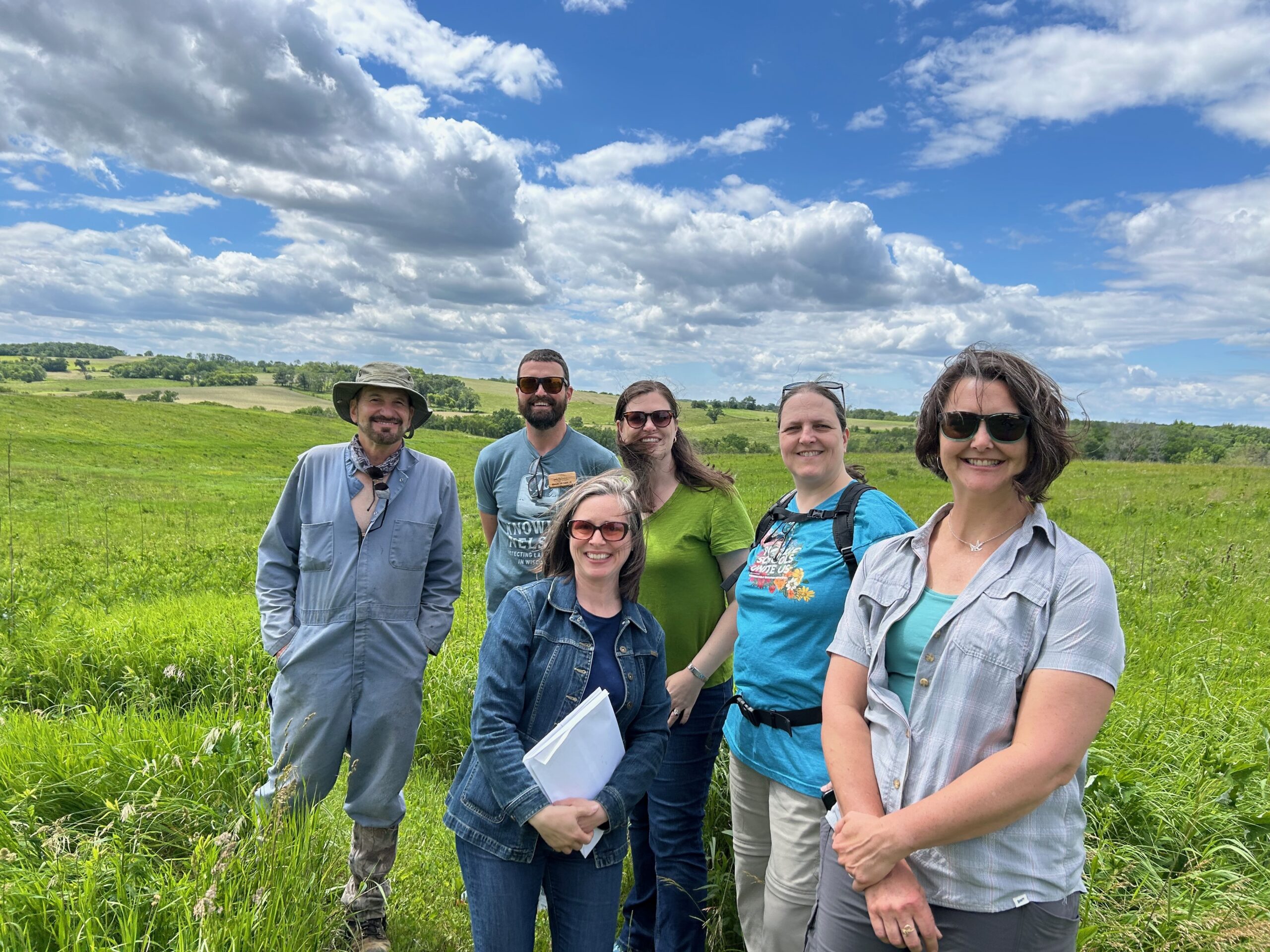 A group of six adults in hiking clothing are standing in an open, green field.