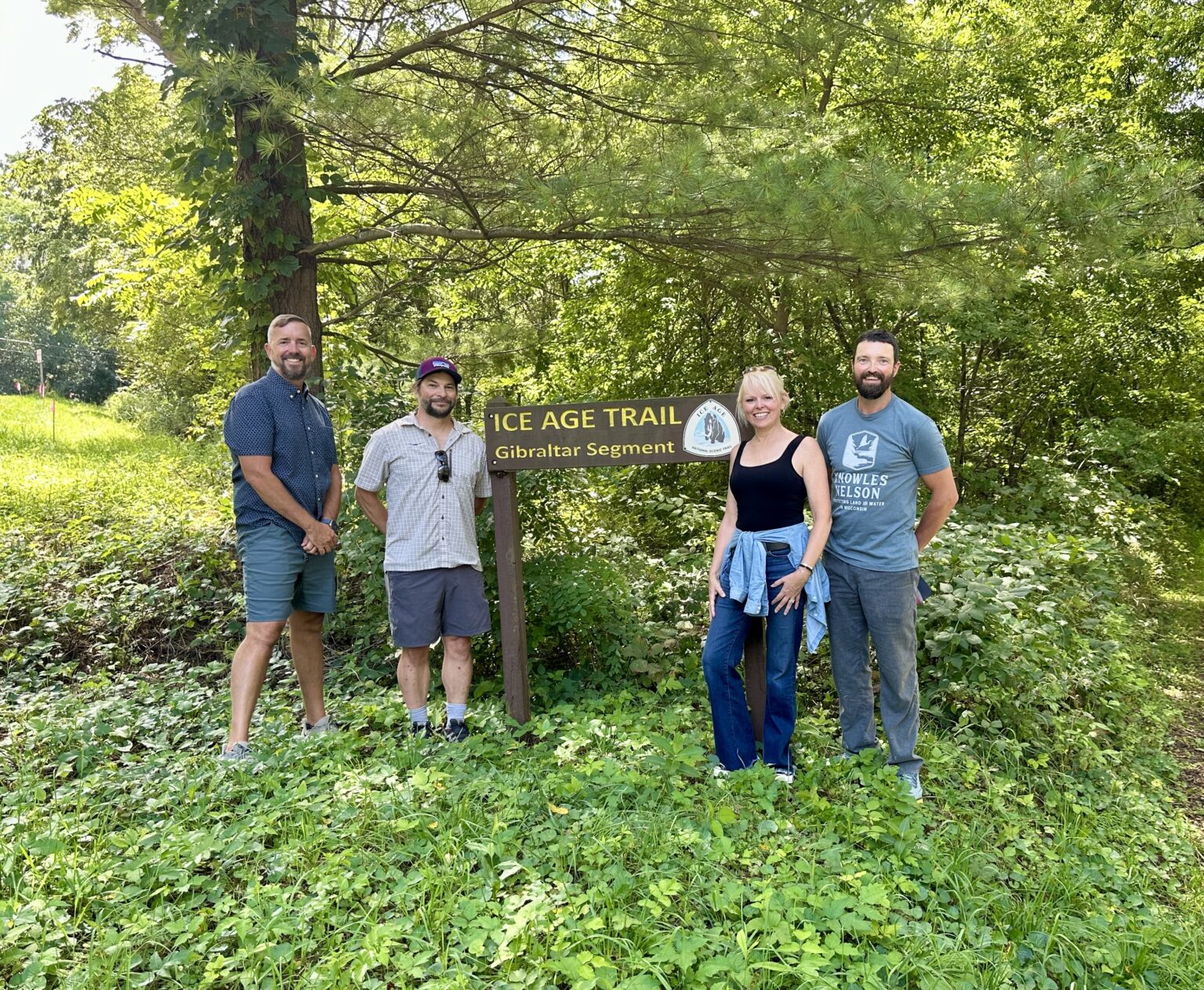 One woman and three men stand near an Ice Age Trail sign in a vegetated area.