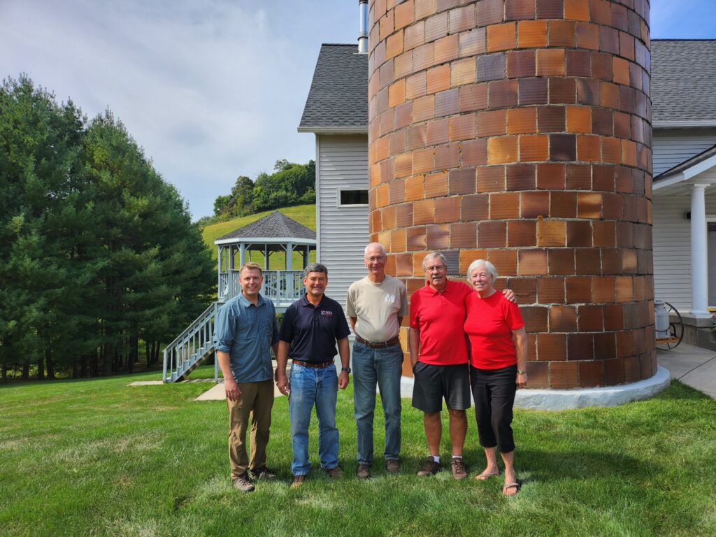 Five adults stand gathered in front of a silo and house.