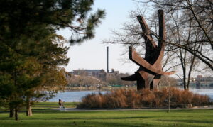 A large metal sculpture is displayed along a riverfront walkway. City buildings are across the riverbank. A child is riding a bike.