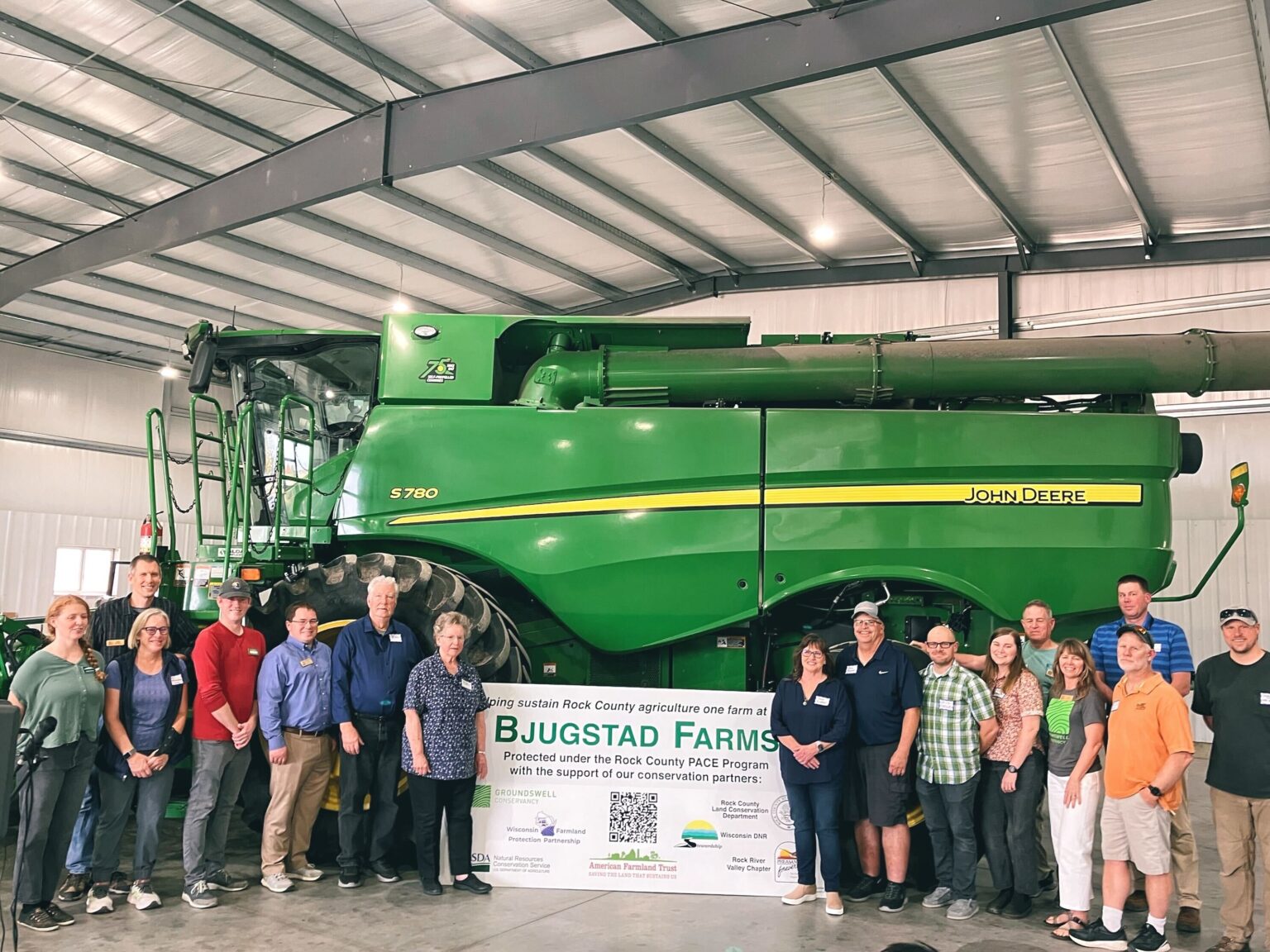 A group of 16 adults standing in front of a large green farming combine. They are indoors, posing for a photo with a sign.