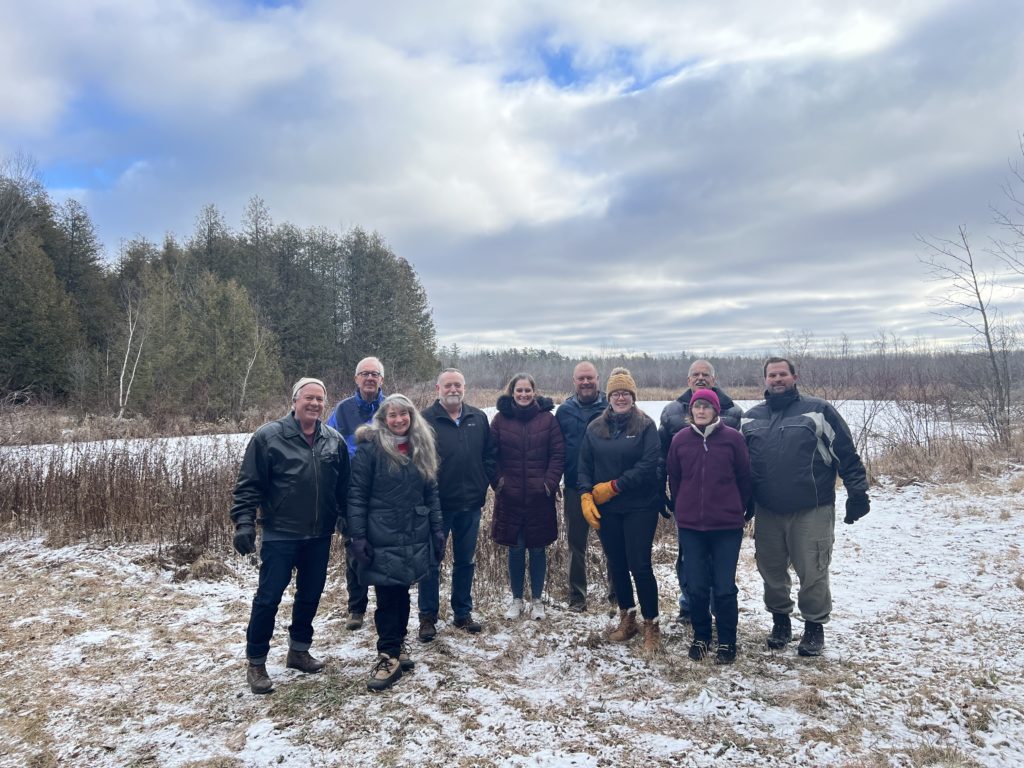 A group of adults standing in an clearing. They are wearing winter clothes and the ground has a light dusting of snow.