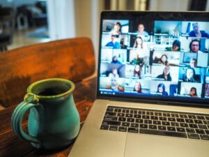 An open laptop on a table displaying a number of people on a Zoom call. There is a green coffee mug next to the laptop.