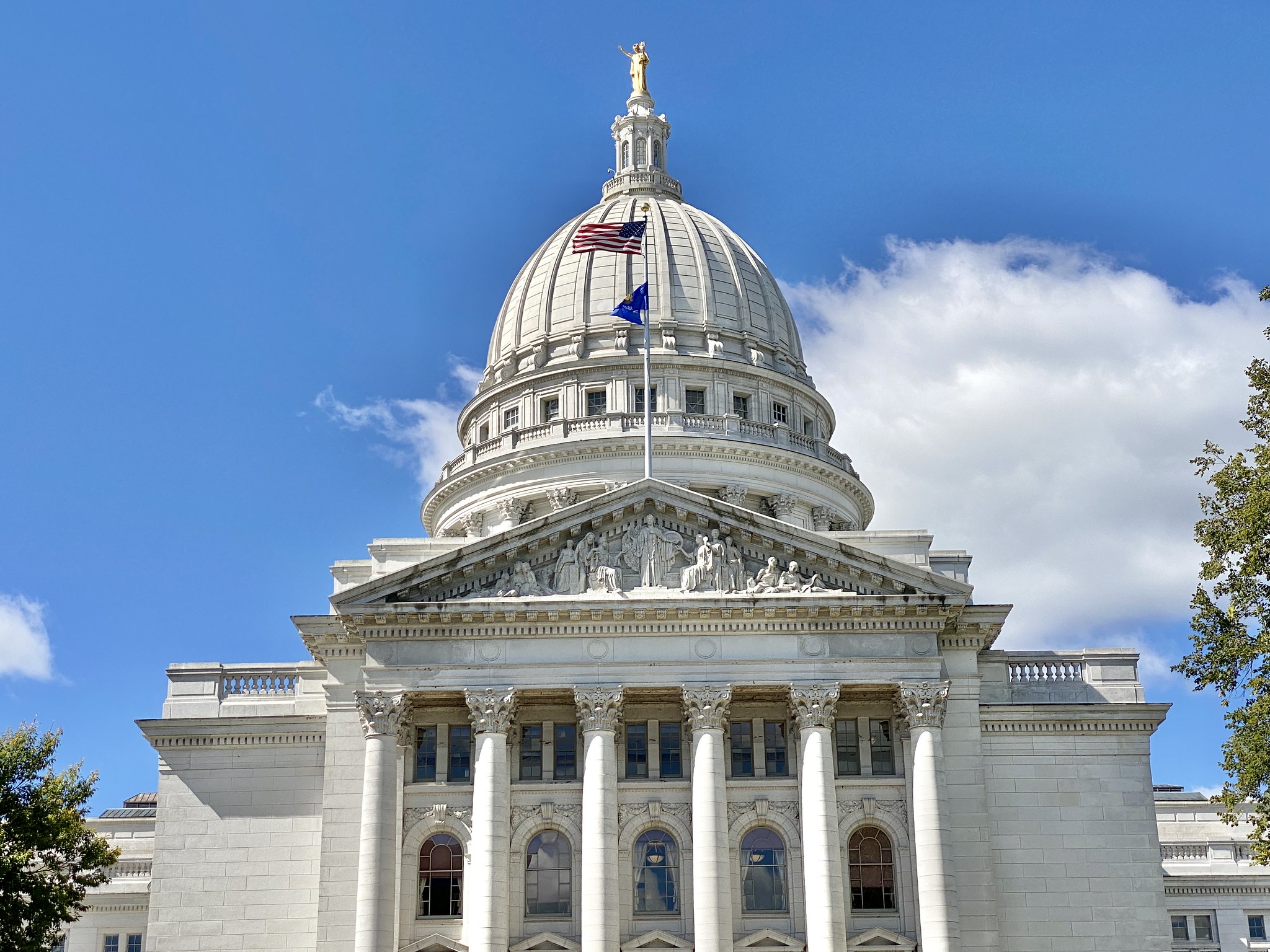 The Wisconsin State Capitol building in Madison, featuring a white granite facade, Corinthian columns, and a prominent dome topped with flags and a statue.