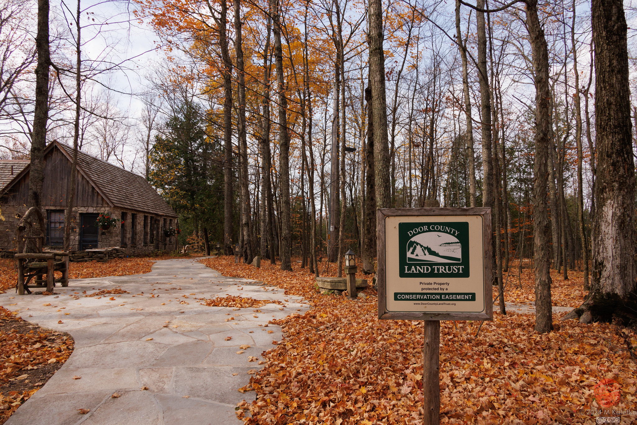 Concrete path leading to a rustic cabin in the woods, with autumn leaves scattered around and a Door County Land Trust conservation easement sign.