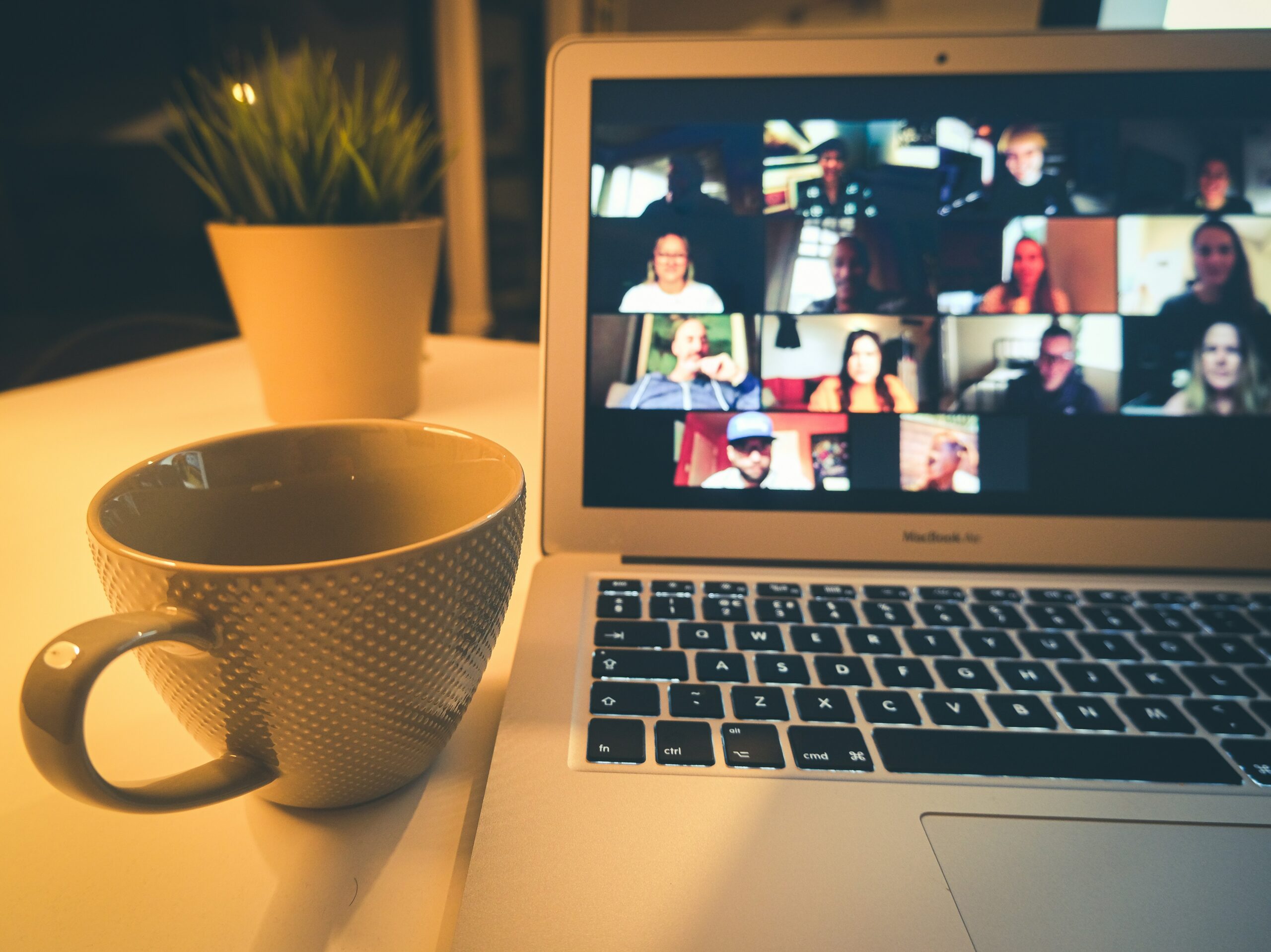 A laptop showing a video call grid, with a coffee mug and plant beside it.