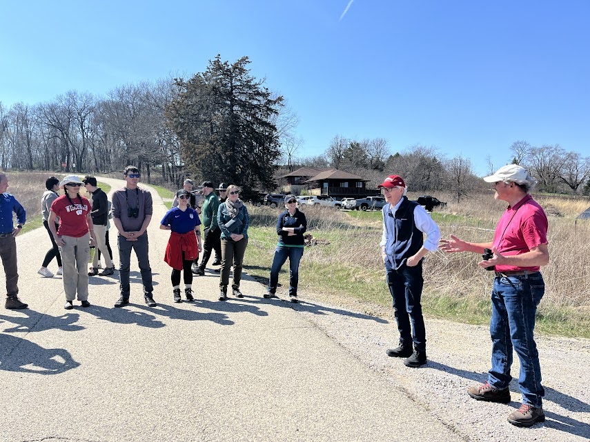 A group of adults in hiking clothing standing outside listening to a man speak.