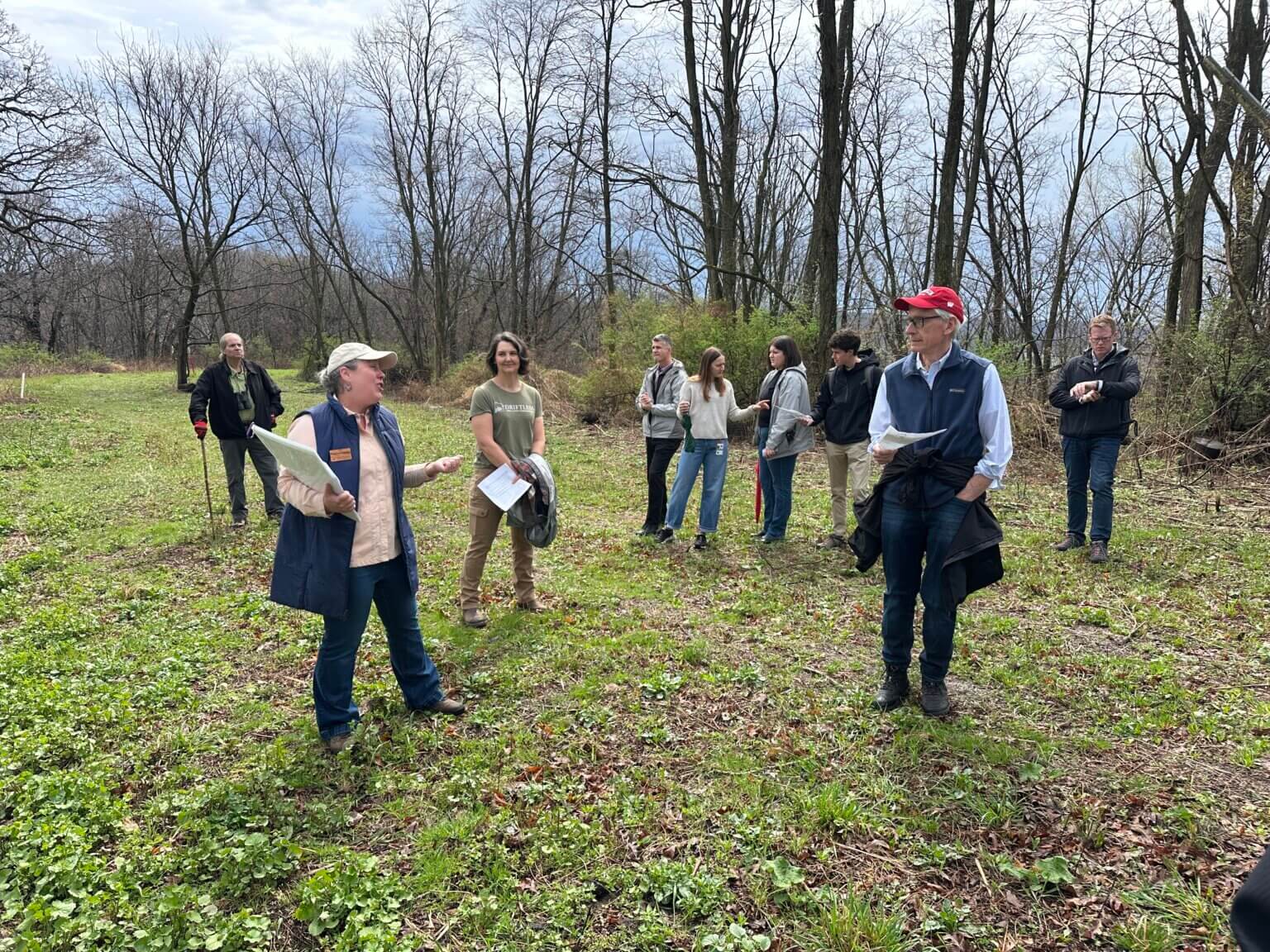 A group of people including Governor Evers (in red cap) explore a wooded area. Spring trees are beginning to bud.