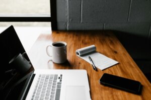An open laptop, coffee mug, and notepad on a wood surface.