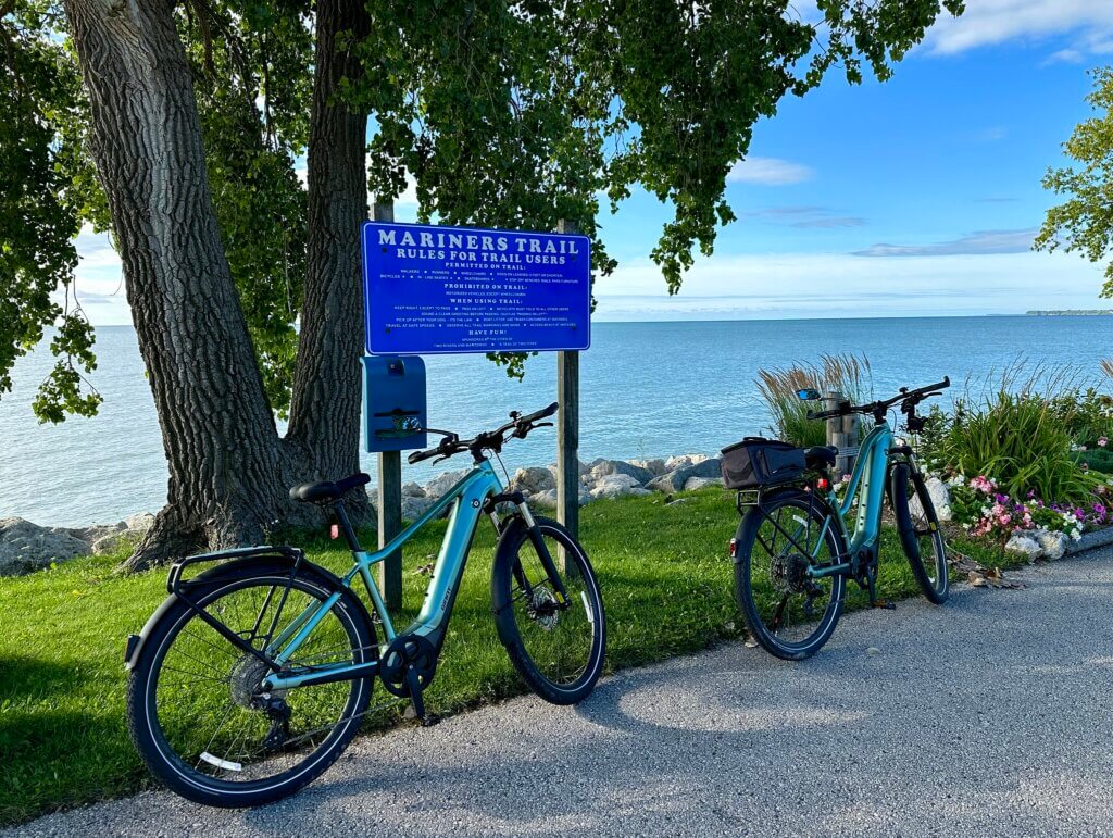 Two light blue e-bikes parked beside a Mariners Trail sign along a scenic waterfront with trees, rocky shoreline, and calm water.