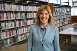 Woman in light blue blazer smiling in library with bookshelves in background.
