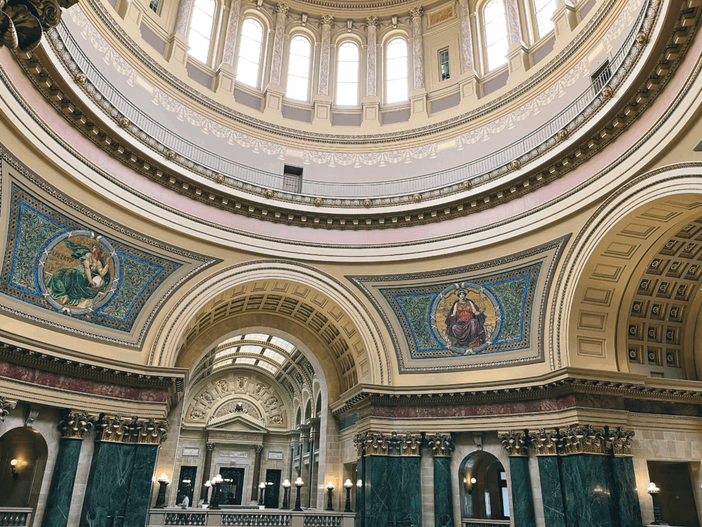 Ornate interior of the Wisconsin Capitol rotunda with a domed ceiling, decorative murals, marble columns, and classical architectural details.