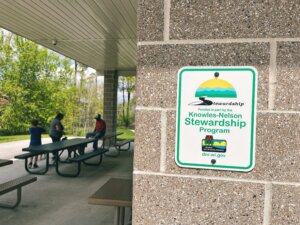 A Knowles-Nelson Stewardship Program sign mounted on a brick wall next to a covered picnic area where people are seated at tables, with green trees visible in the background on a sunny day.