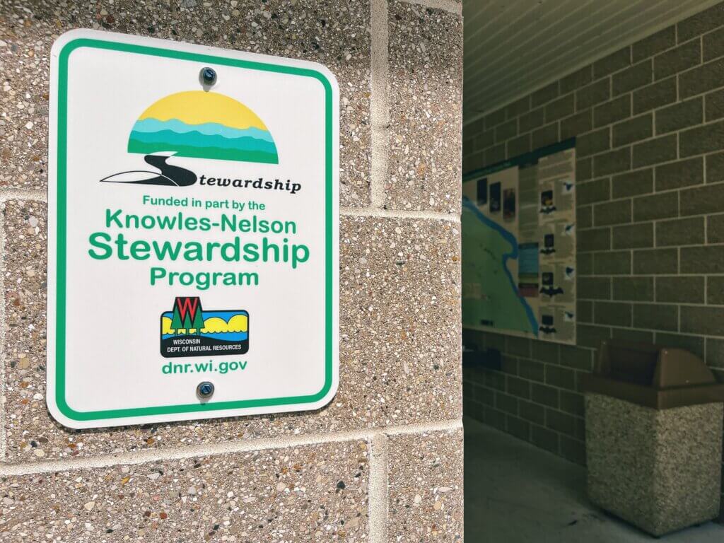 A white sign with green border mounted on a concrete wall displays the Knowles-Nelson Stewardship Program logo, with the Wisconsin Department of Natural Resources logo and website below.