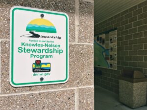 A white sign with green border mounted on a concrete wall displays the Knowles-Nelson Stewardship Program logo, with the Wisconsin Department of Natural Resources logo and website below.