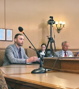 A man in a gray suit seated at conference table with microphone during legislative hearing, with two people visible in the background.