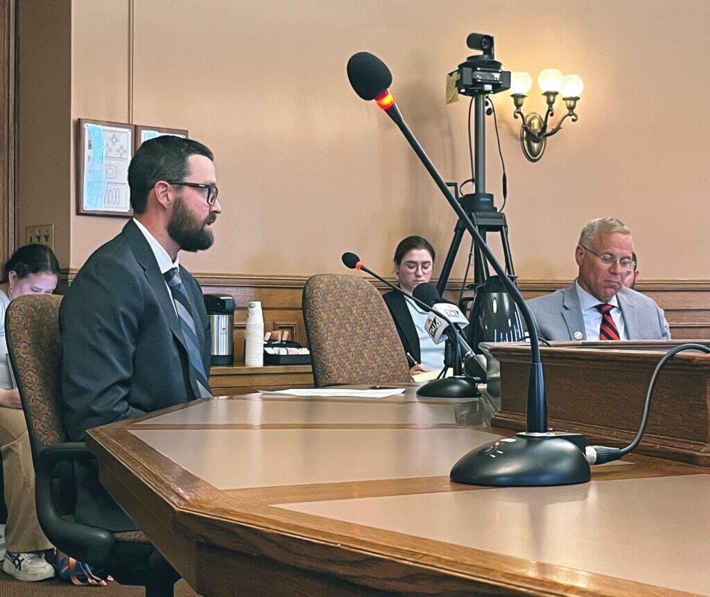 A man in a dark suit and glasses testifies at a microphone in a formal legislative hearing room, with committee members seated at a curved wooden desk in the background and recording equipment visible.