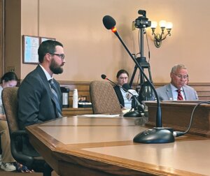 A man in a dark suit and glasses testifies at a microphone in a formal legislative hearing room, with committee members seated at a curved wooden desk in the background and recording equipment visible.