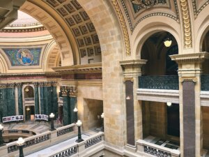 Interior view of the Wisconsin State Capitol rotunda showing ornate architecture with detailed stone arches, decorative moldings, and classical columns with Corinthian capitals, along with green marble wainscoting and a partial view of the upper gallery levels.
