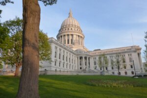 Wisconsin State Capitol building with its distinctive white dome and neoclassical architecture, viewed from the surrounding lawn with a large tree in the foreground.