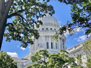 The Wisconsin State Capitol building with its distinctive white dome, framed by green tree branches against a blue sky with white clouds.