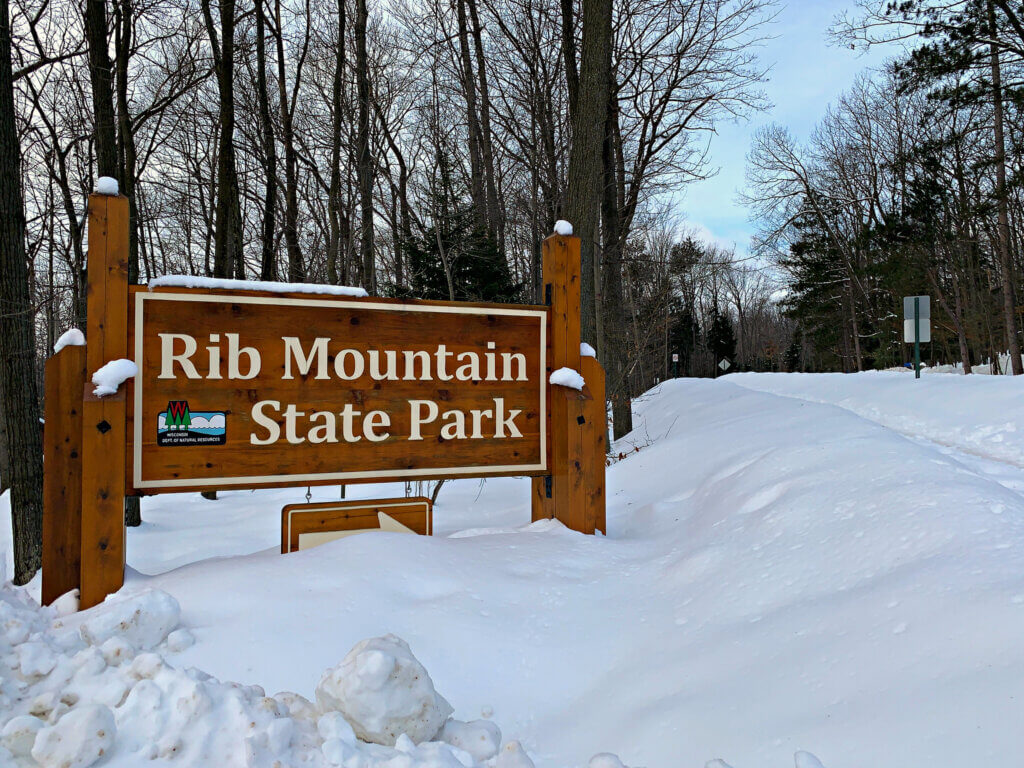 Snow-covered entrance sign for Rib Mountain State Park, surrounded by bare winter trees.