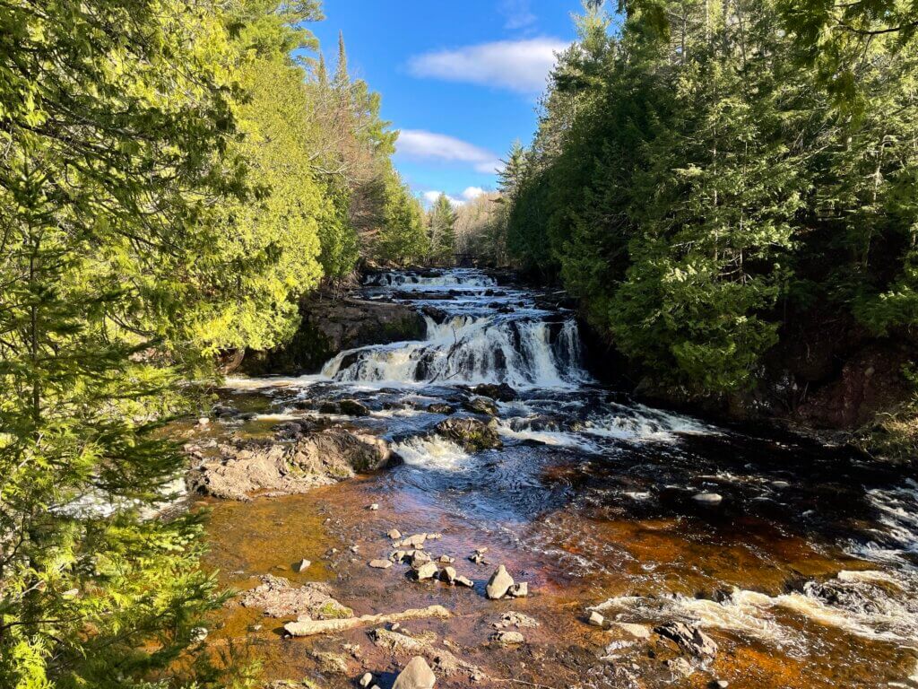 A wide, multi-tiered waterfall flowing over rocky ledges, surrounded by evergreen trees under a bright blue sky.