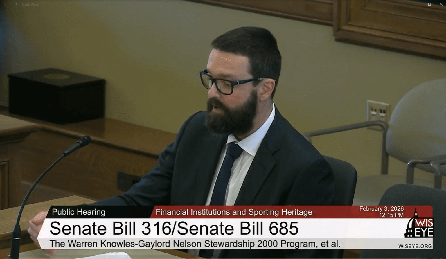 Man in a suit and glasses testifies at a Wisconsin Senate committee hearing on Knowles-Nelson stewardship legislation, with WisconsinEye broadcast graphics visible on screen.