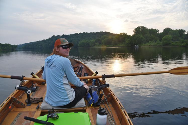 Person rowing a small wooden boat across a calm river at sunset, with tree-lined hills and reflections on the water.