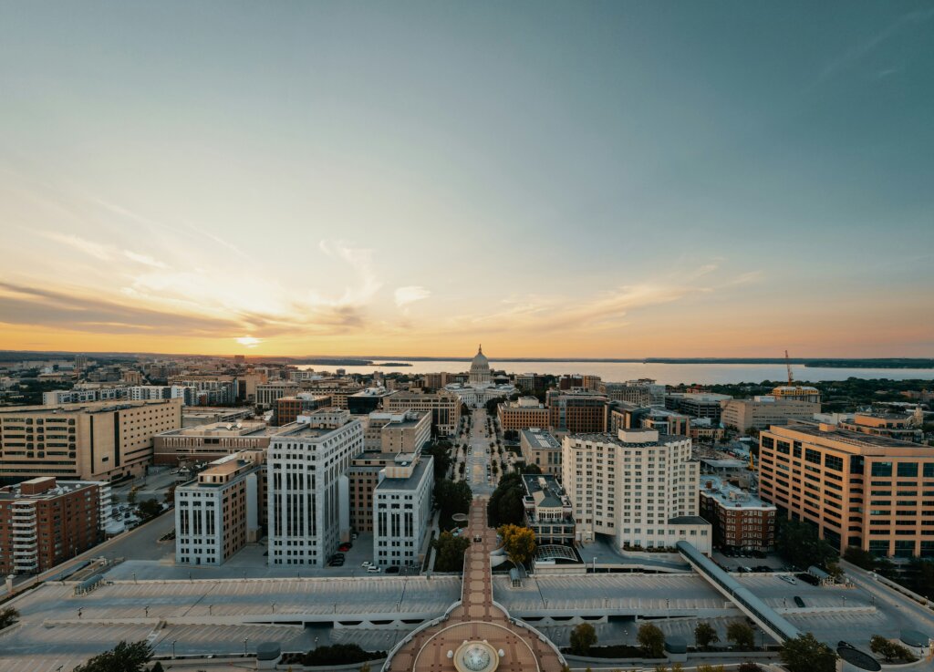 Aerial view of downtown Madison, Wisconsin at sunset, with the Wisconsin State Capitol centered along State Street and Lake Mendota in the background.