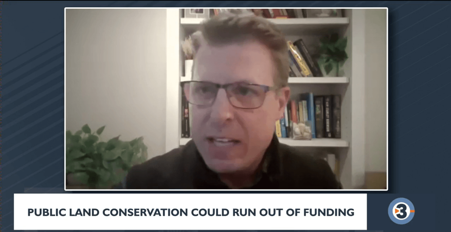 A man wearing glasses speaks during a virtual interview, with a bookshelf behind him, while on-screen text reads “Public land conservation could run out of funding.”