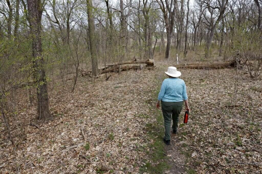A person wearing a white sun hat and teal shirt walks along a woodland trail through a bare early-spring forest, carrying a red water bottle.