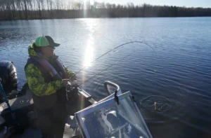 A person in a green & yellow camo jacket and cap reels in a fish from a motorboat on a calm northern Wisconsin lake, with sunlight glinting off the water and bare trees lining the far shore.