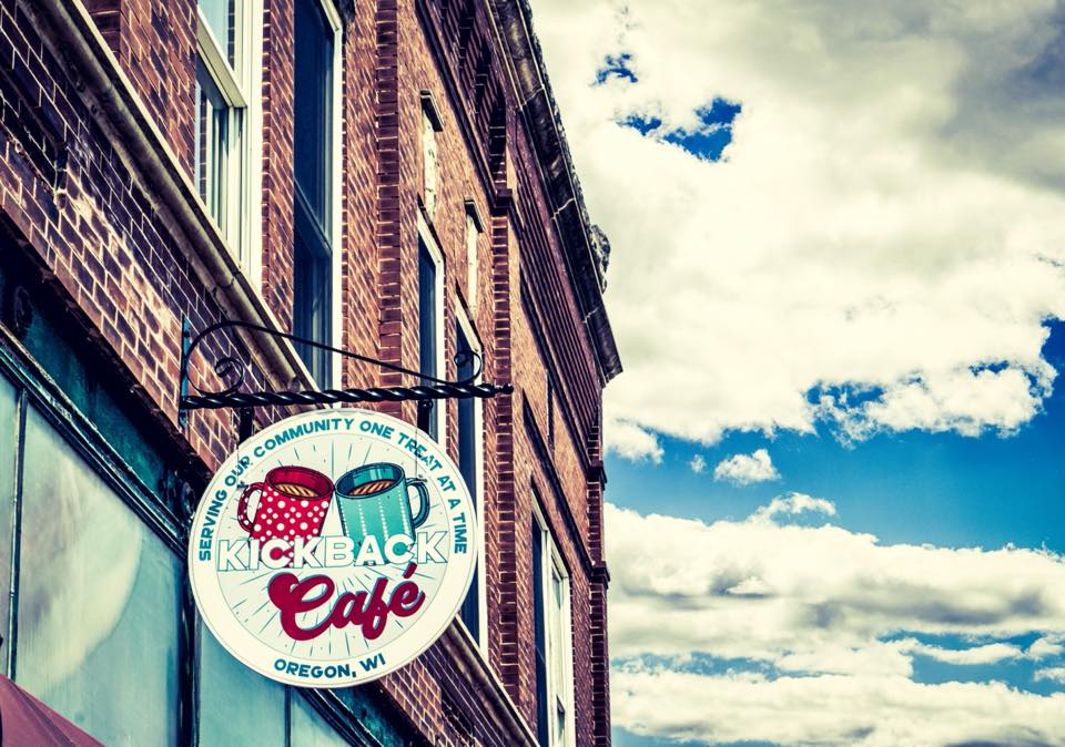 Image 1: A circular hanging sign for Kickback Café in Oregon, Wisconsin reads "Serving our community one treat at a time," mounted on a red brick building against a partly cloudy sky.